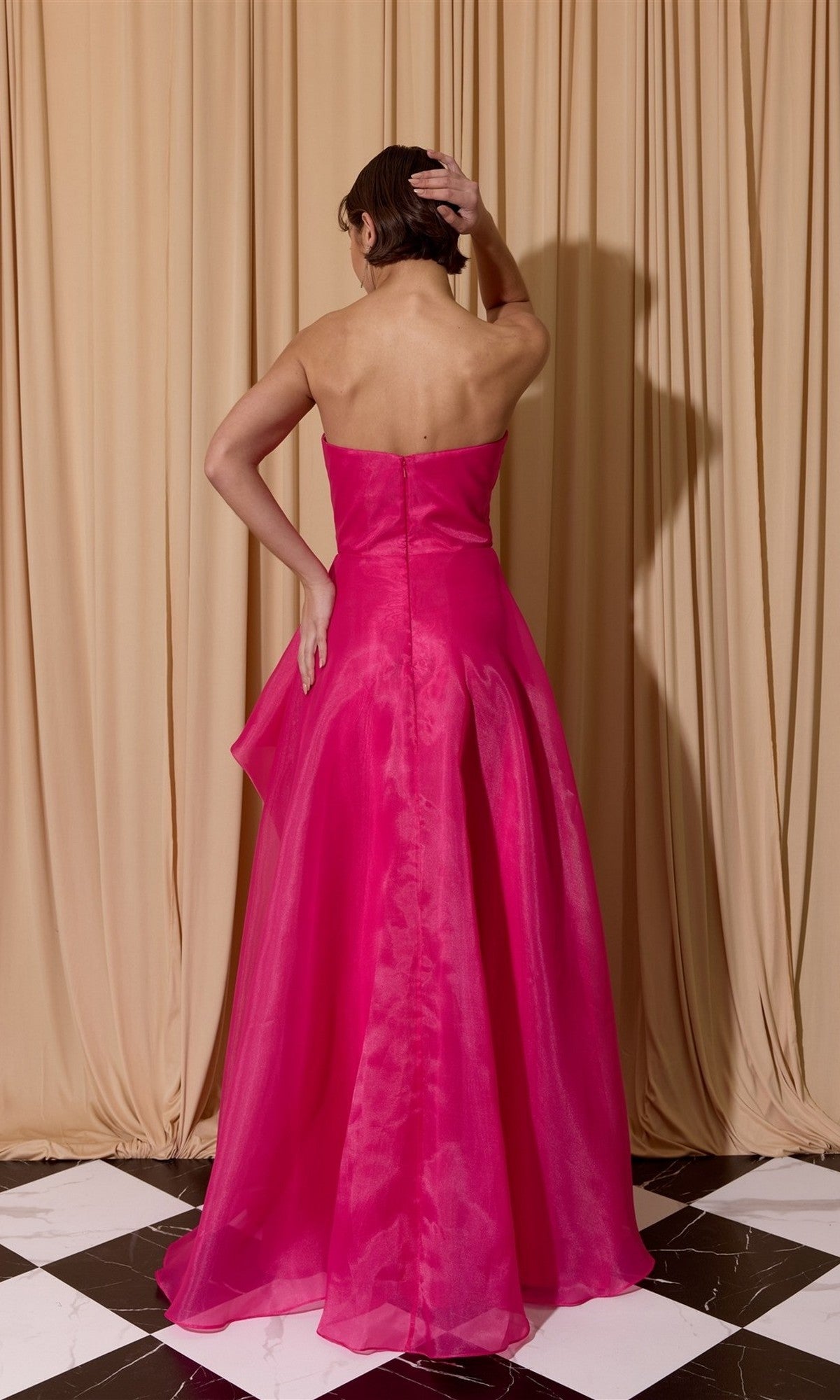 A woman stands on a black and white tiled floor, facing away, wearing the bright pink Eureka EK206 Long Prom Dress in a ball gown style before beige curtains—an elegant fashion moment.